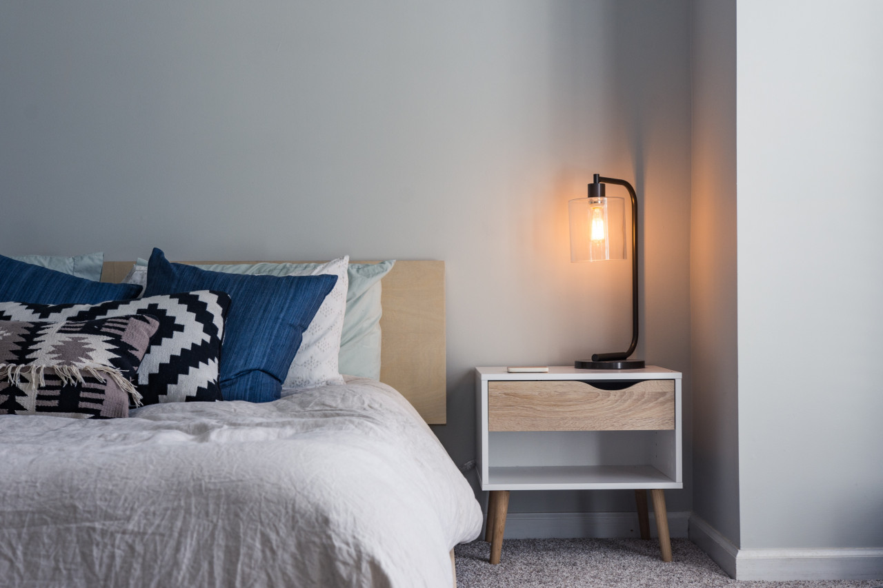A made bed and nightstand with a lamp turned on.