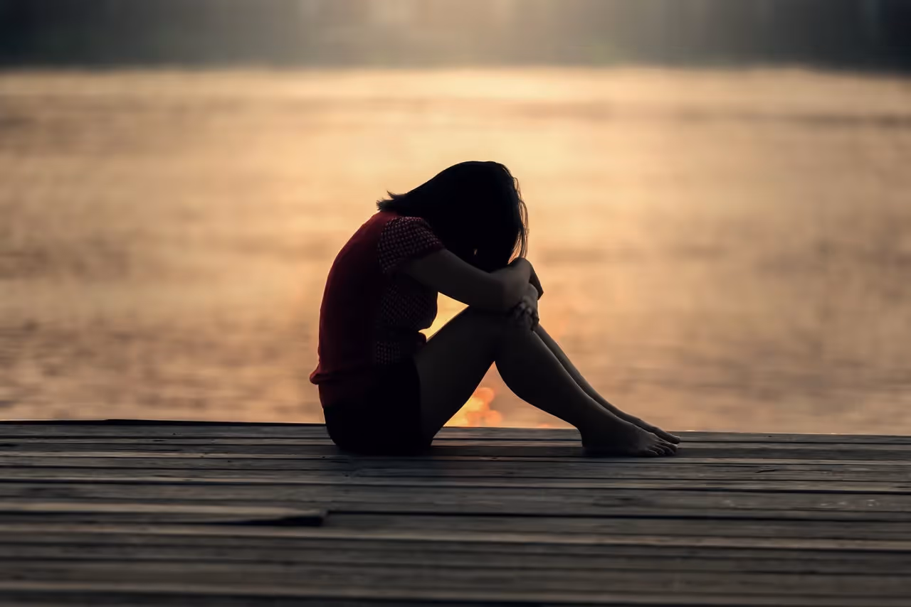 Woman sitting on a dock near a body of water resting her arms on her knees and holding her head down.