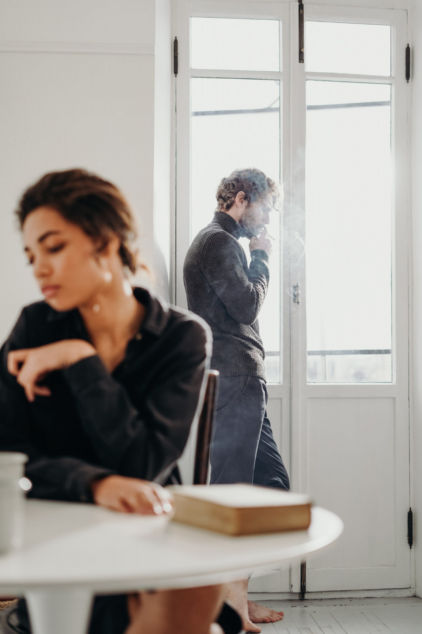 Woman sitting at table with a man standing at a door behind her smoking