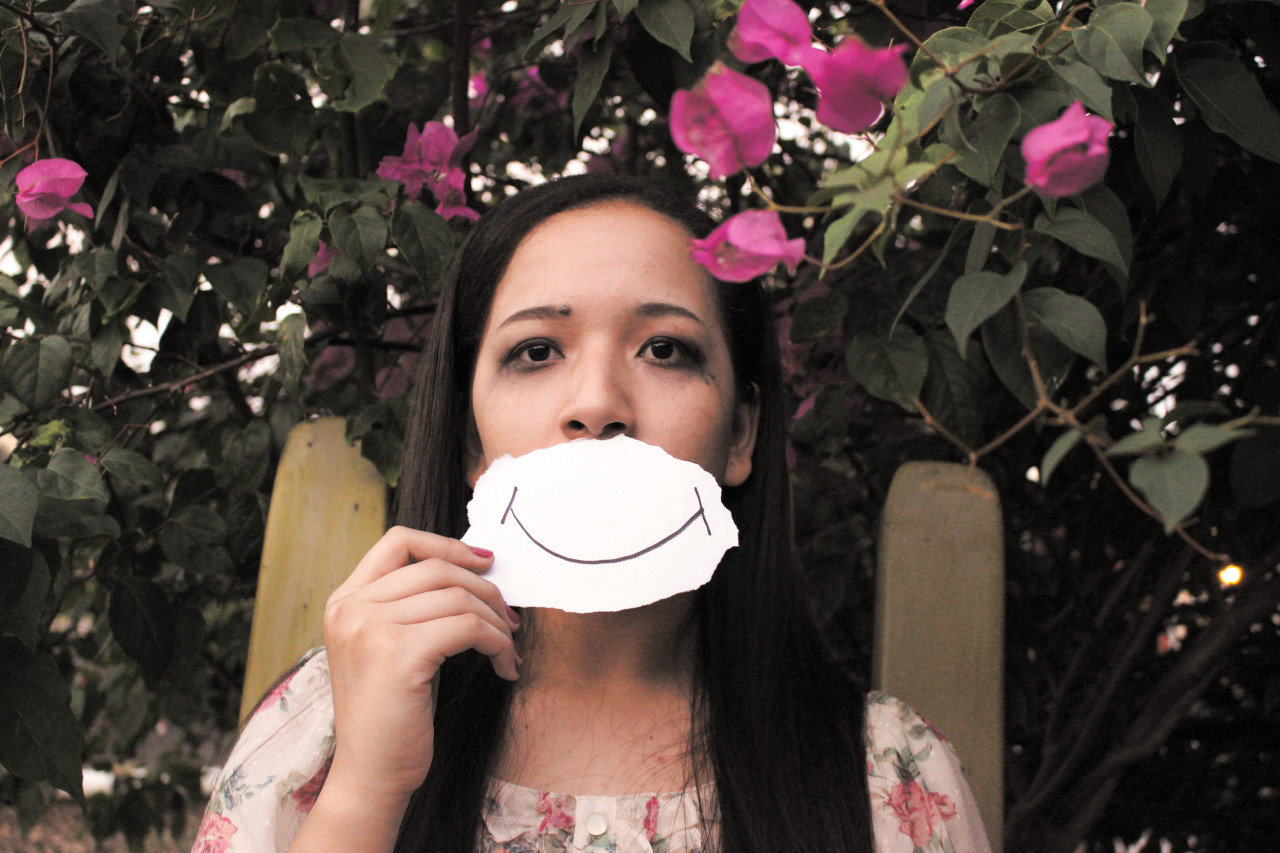 a teen with smeared eye makeup holding a piece of paper with a smile drawn on it over her mouth.