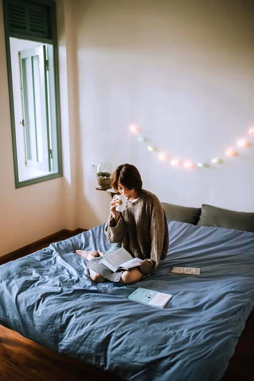 A woman reading a book and drinking coffee on her bed.