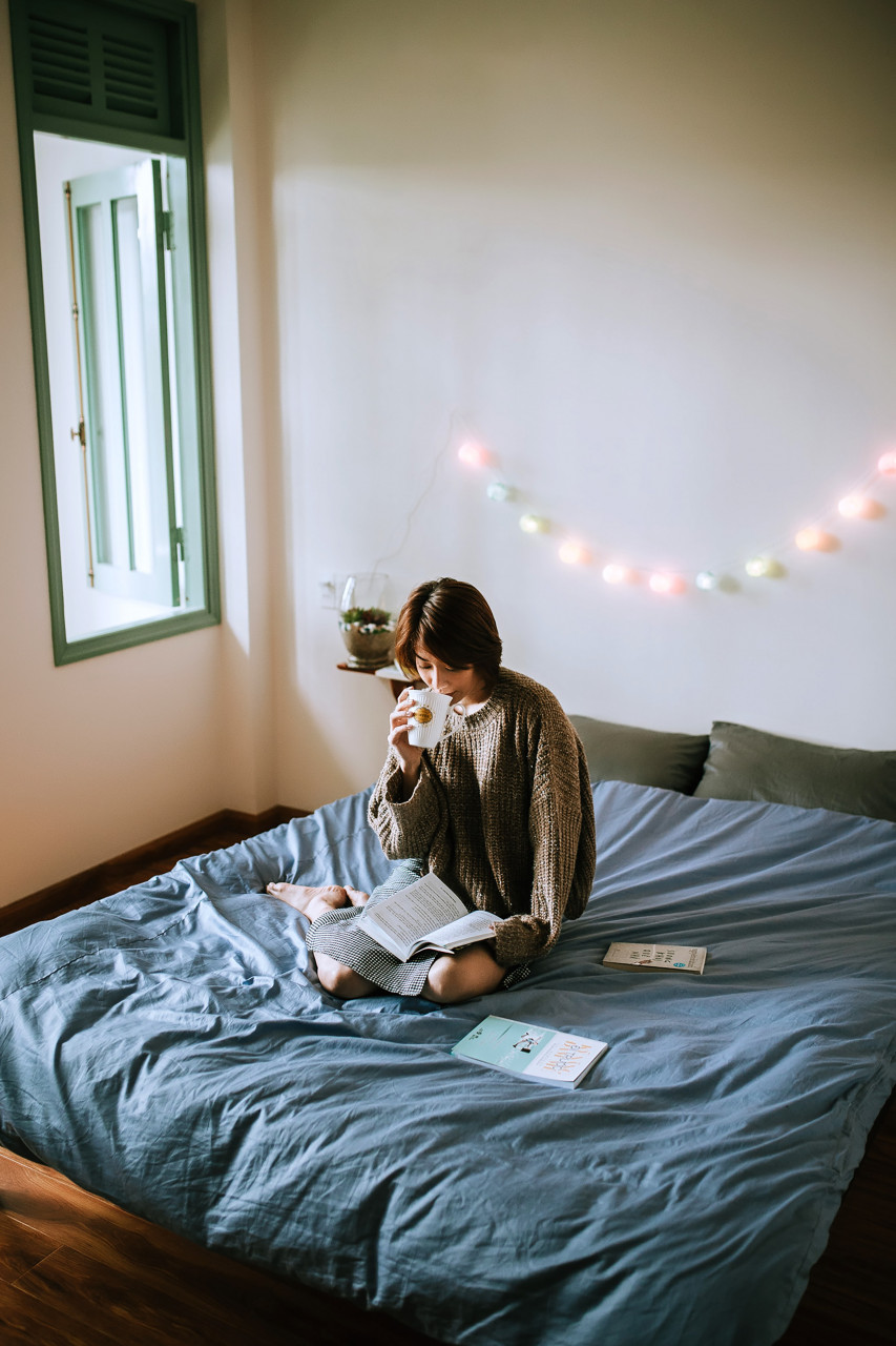A woman reading a book and drinking coffee on her bed.