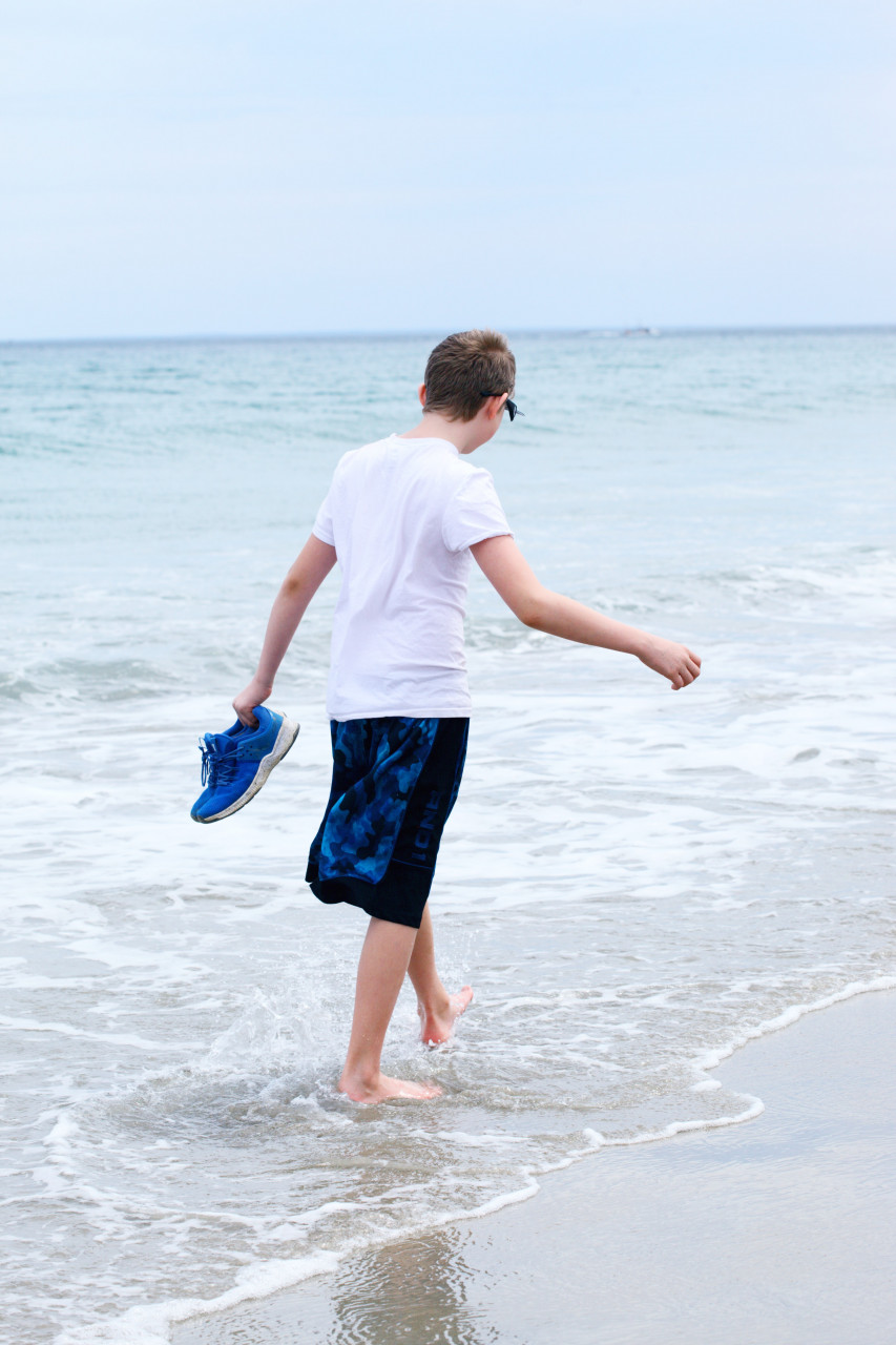 A boy holding a blue shoe while walking on the beach