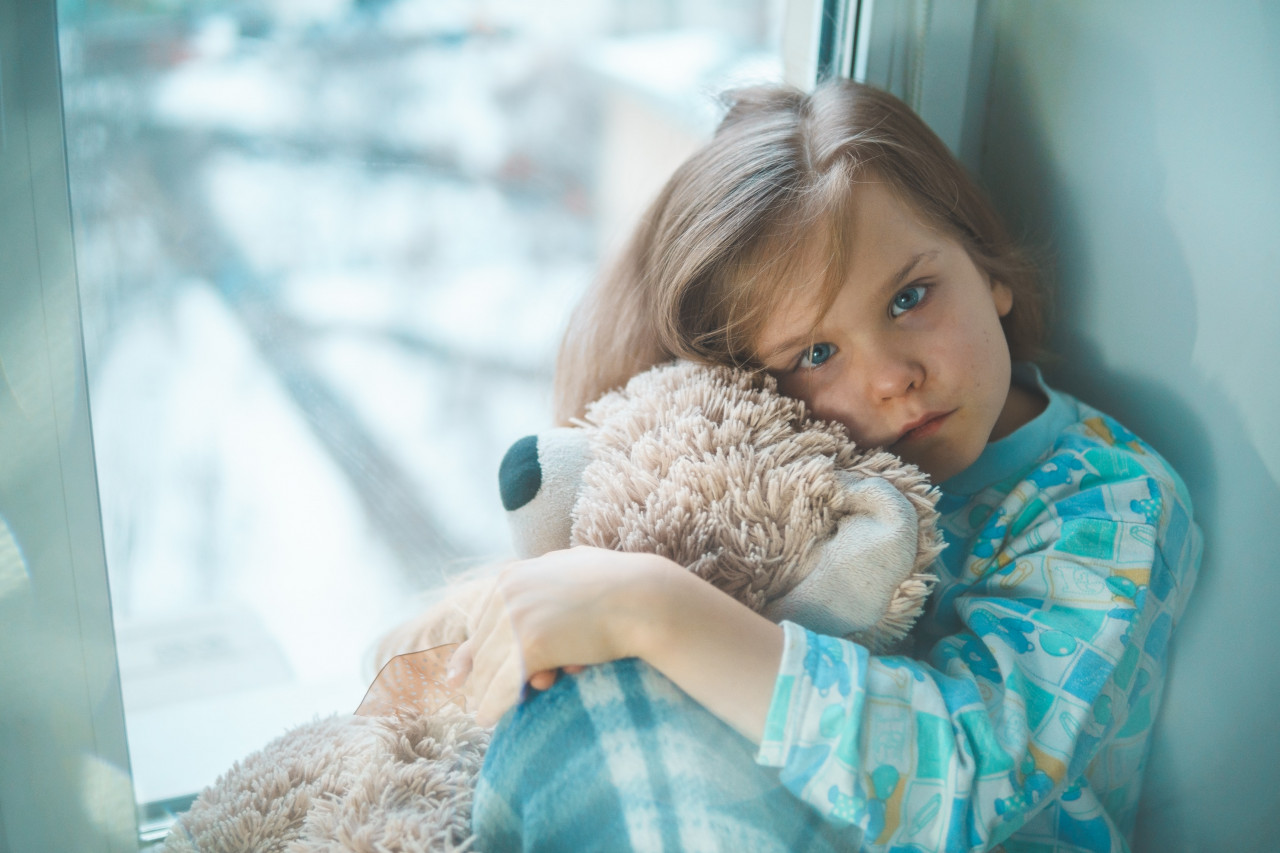 A child sitting next to a window holding a teddy bear