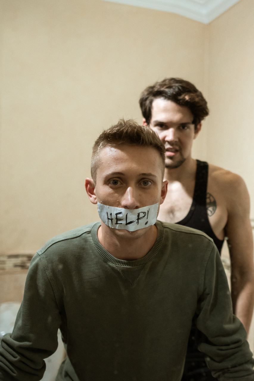 A man standing behind another man who has tape with the word Help written on it over his mouth.