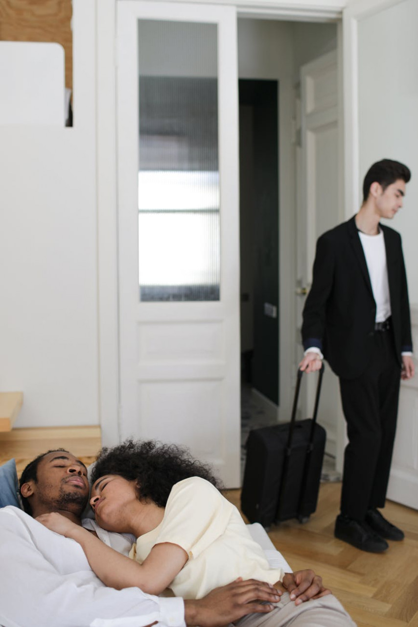 A couple snuggling in bed while a man stands in the doorway with his suitcase.