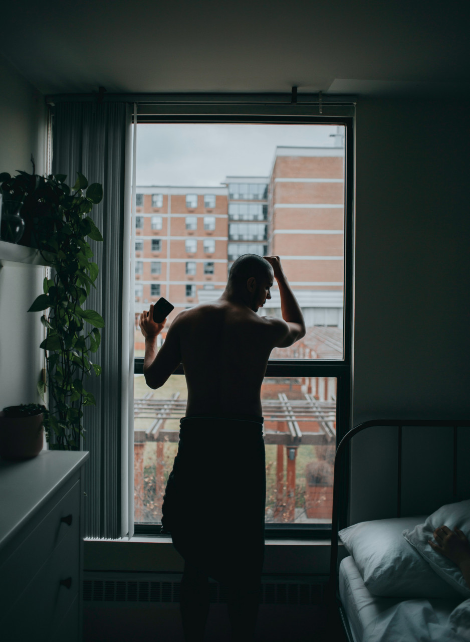A man with his arms resting on the window and his phone in his hands as he looks down on his bed.