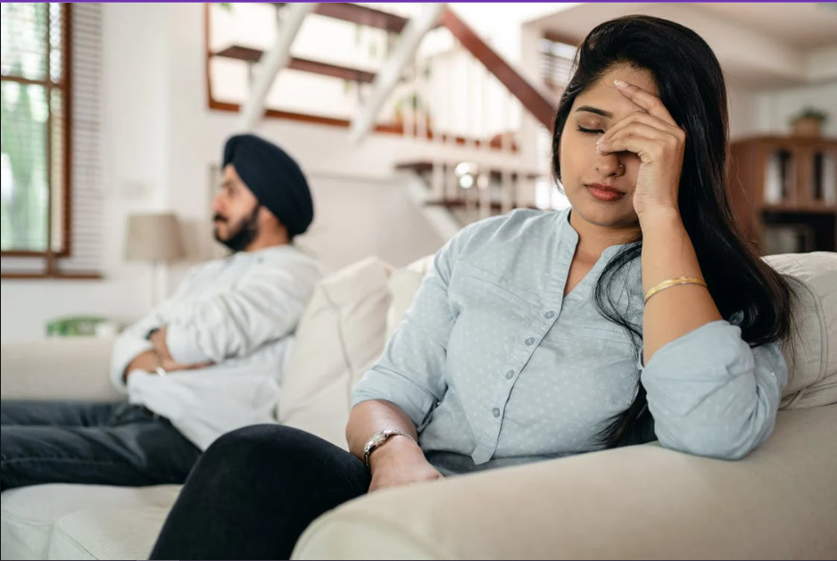 2 people sitting on a couch. The man is ignoring the woman who has her hand on her forehead.