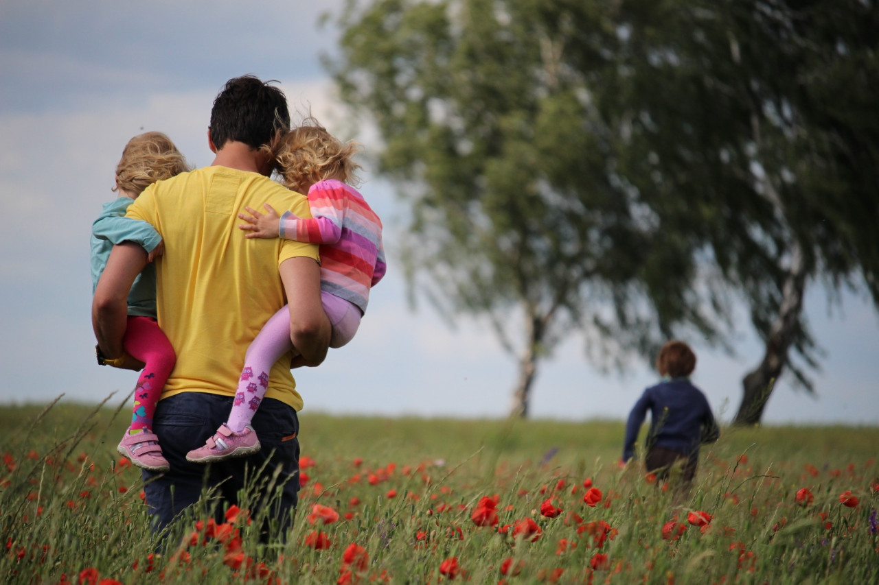 A dad carrying two of his kids while one walks in front.