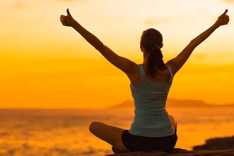 A woman overlooking the water with her arms raised in the air giving thumbs up