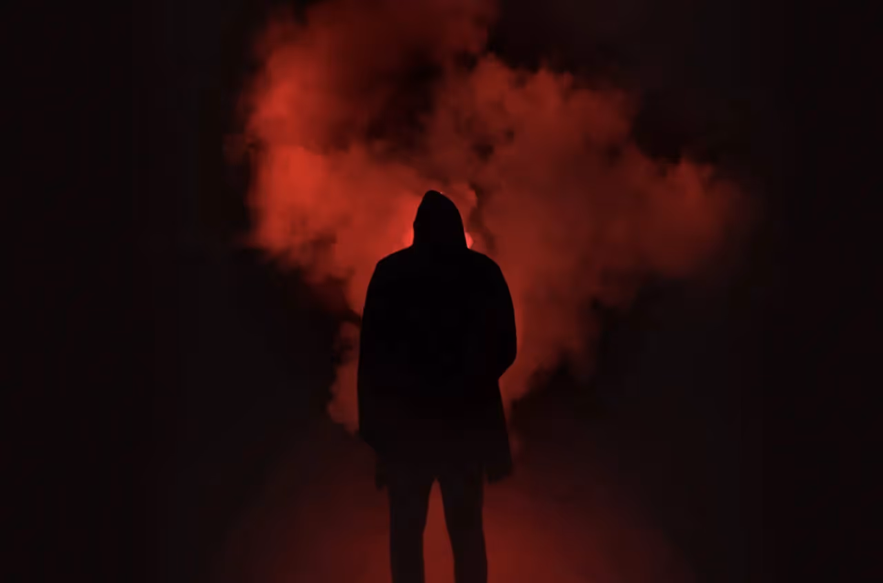 Man wearing dark clothing standing in front of a cloud of smoke lit with a red light.