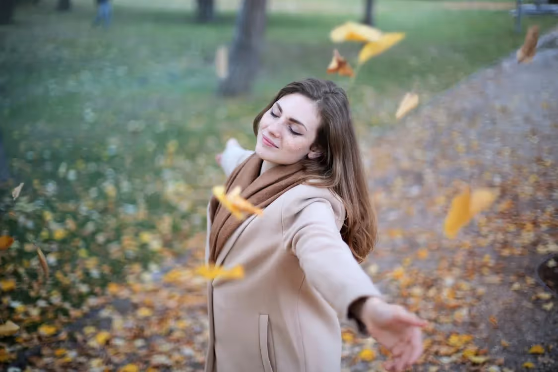 woman in tan coat smiling in falling leaves