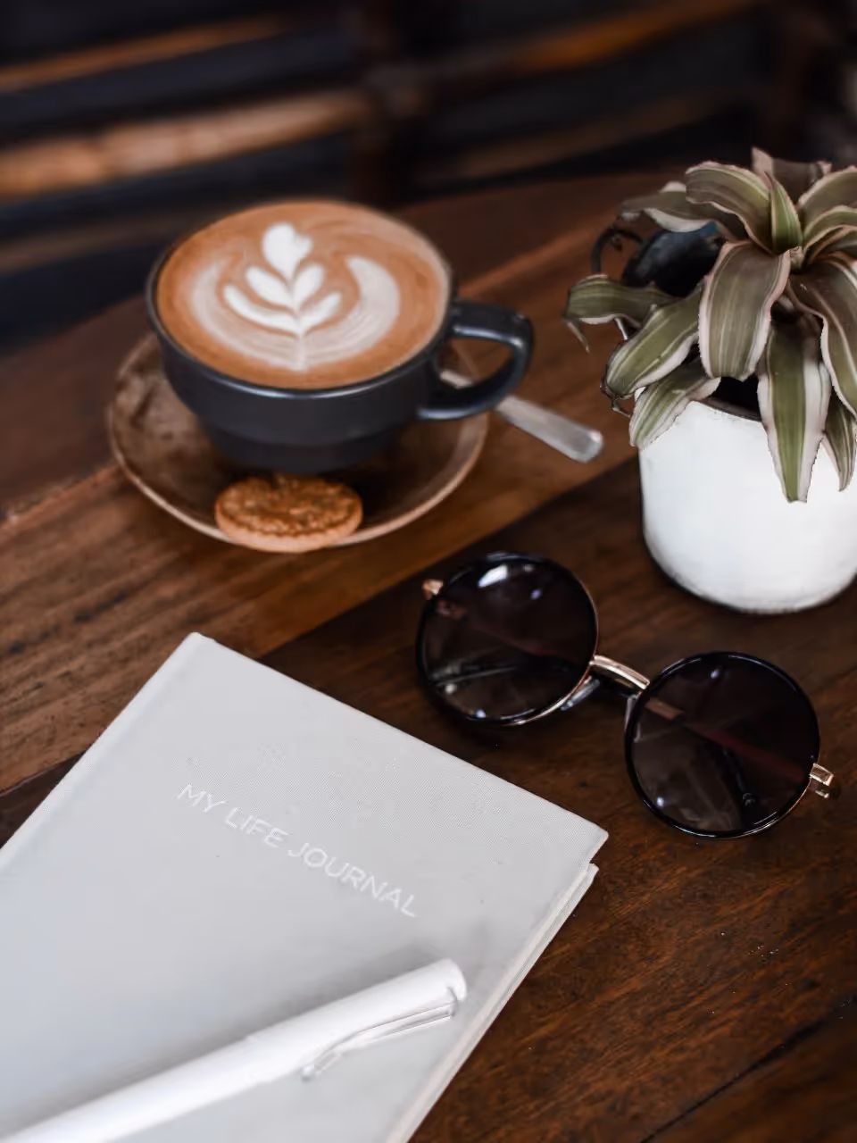 A life journal and white pen sitting on a table next to sunglasses, a plant, and a drink.