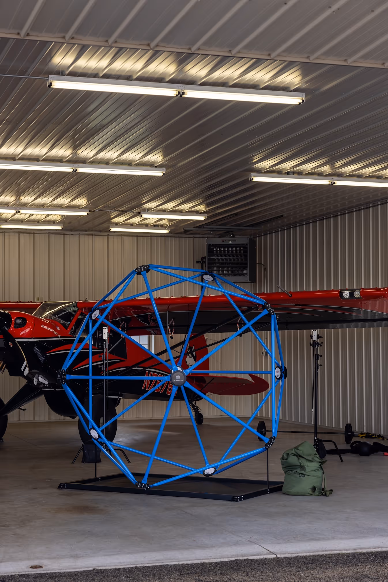 Red and black small airplane inside a metal hangar with a large blue geometric frame structure in front of it.