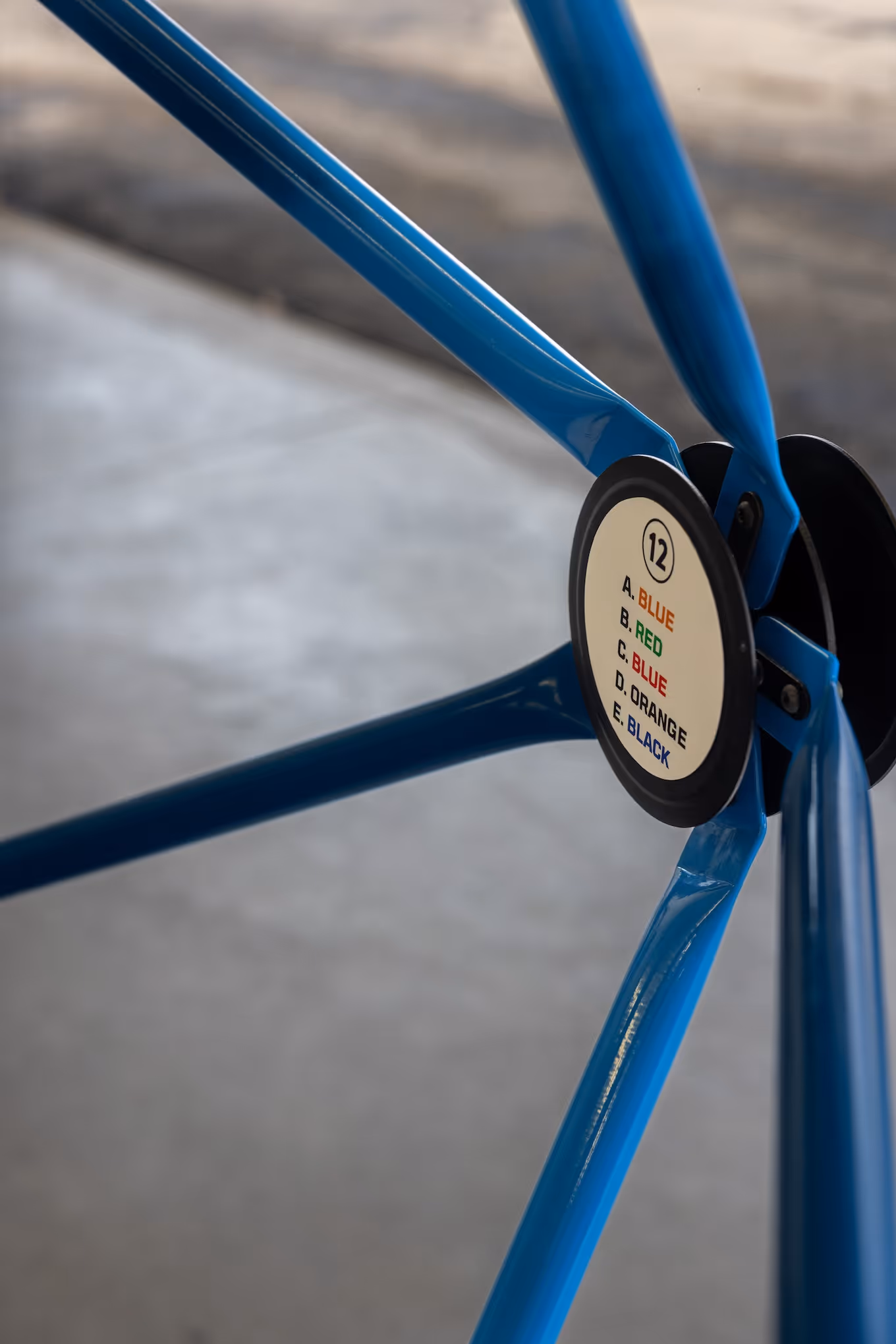 Close-up of a blue metal tripod structure with a circular label showing color-coded options: A. BLUE, B. RED, C. BLUE, D. ORANGE, E. BLACK, numbered 12.