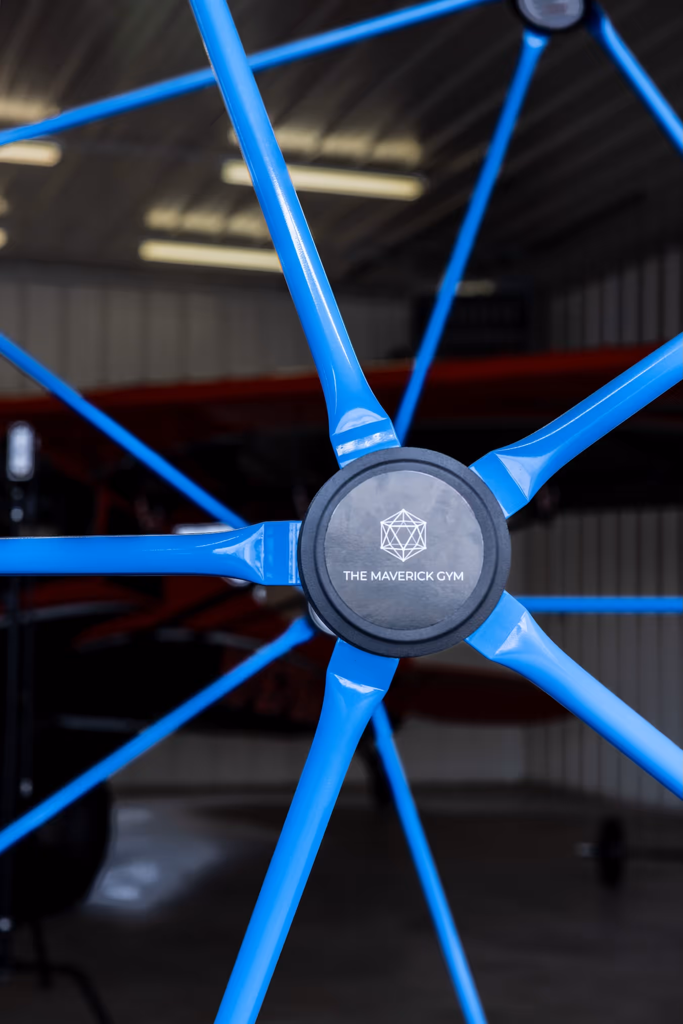Close-up of a blue wheel hub with spokes inside a gym, featuring The Maverick Gym logo in the center.