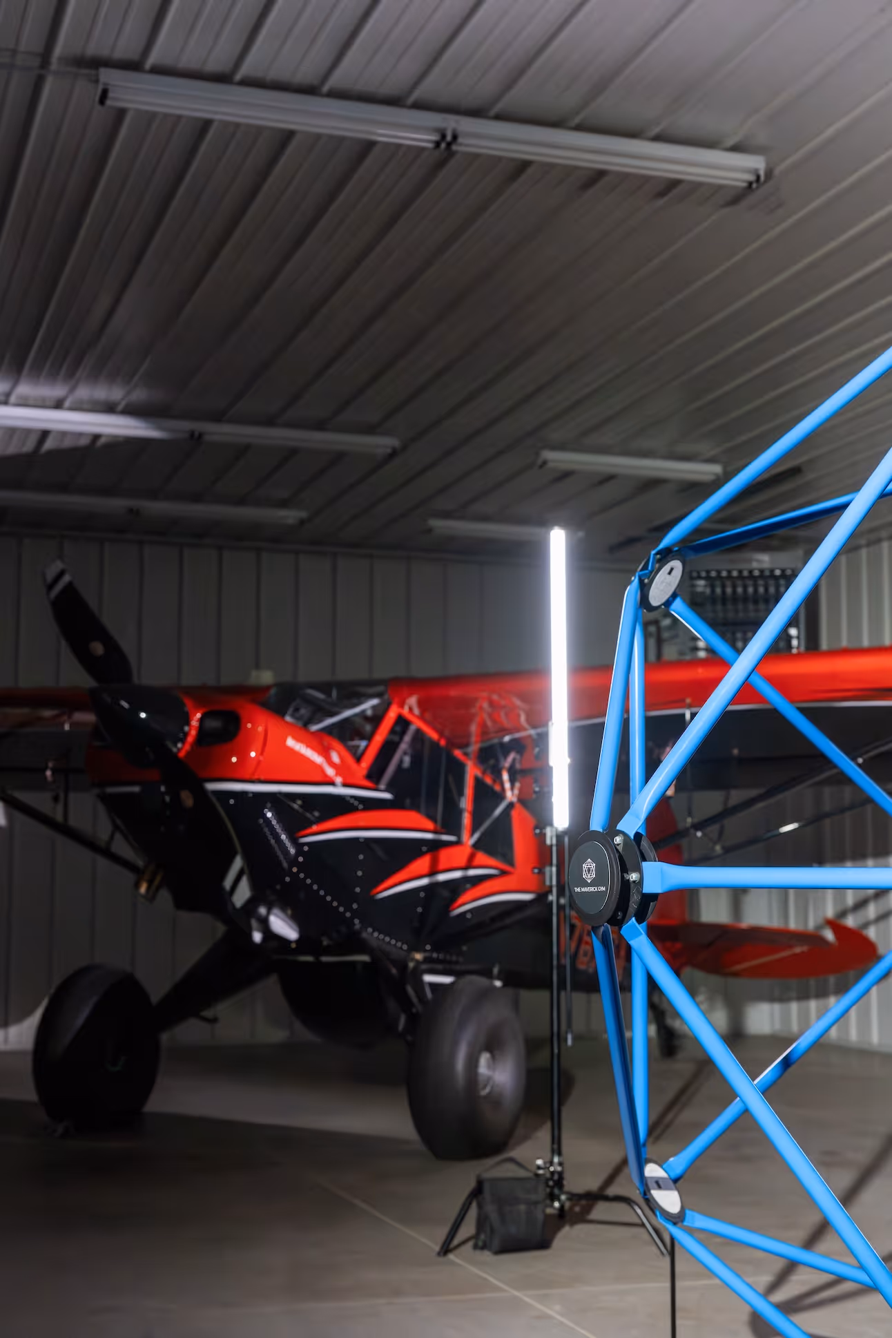 Red and black small aircraft inside a hangar with a bright vertical light and a blue geometric frame structure in the foreground a maverick gym.