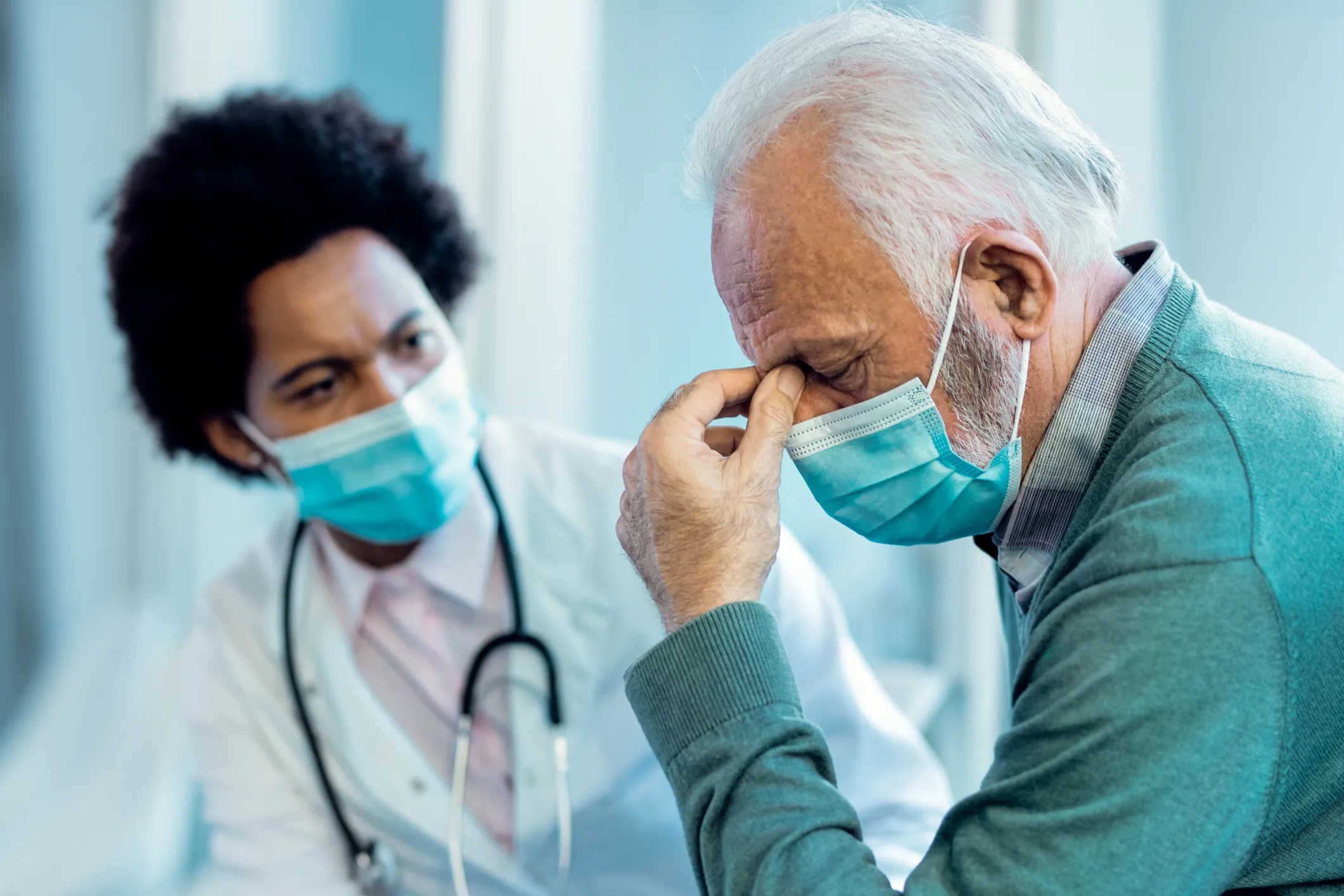 An elderly man wearing a medical mask bows his head while a concerned doctor looks on