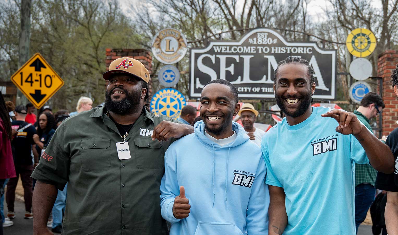Three Black men smile and pose together in front of the Welcome to Historic Selma sign in Selma, Alabama. Two wear Black Male Initiative (BMI) t-shirts; the man in the center gives a thumbs up.