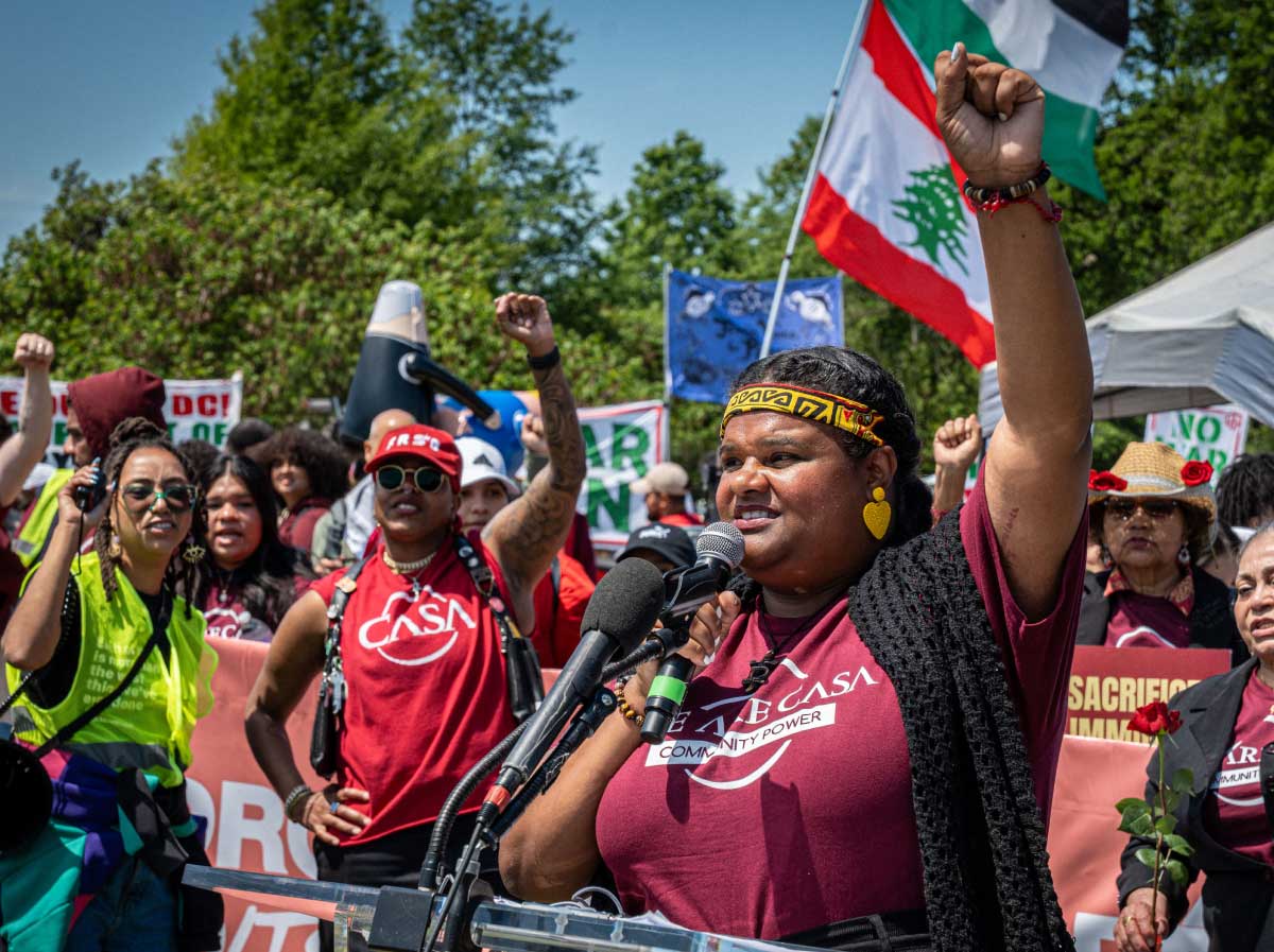 A Black woman in a maroon t-shirt speaks passionately at a microphone podium at an outdoor rally, her fist raised in the air. Behind her a crowd of protesters wear CASA t-shirts and hold signs, with a Lebanese flag visible overhead.