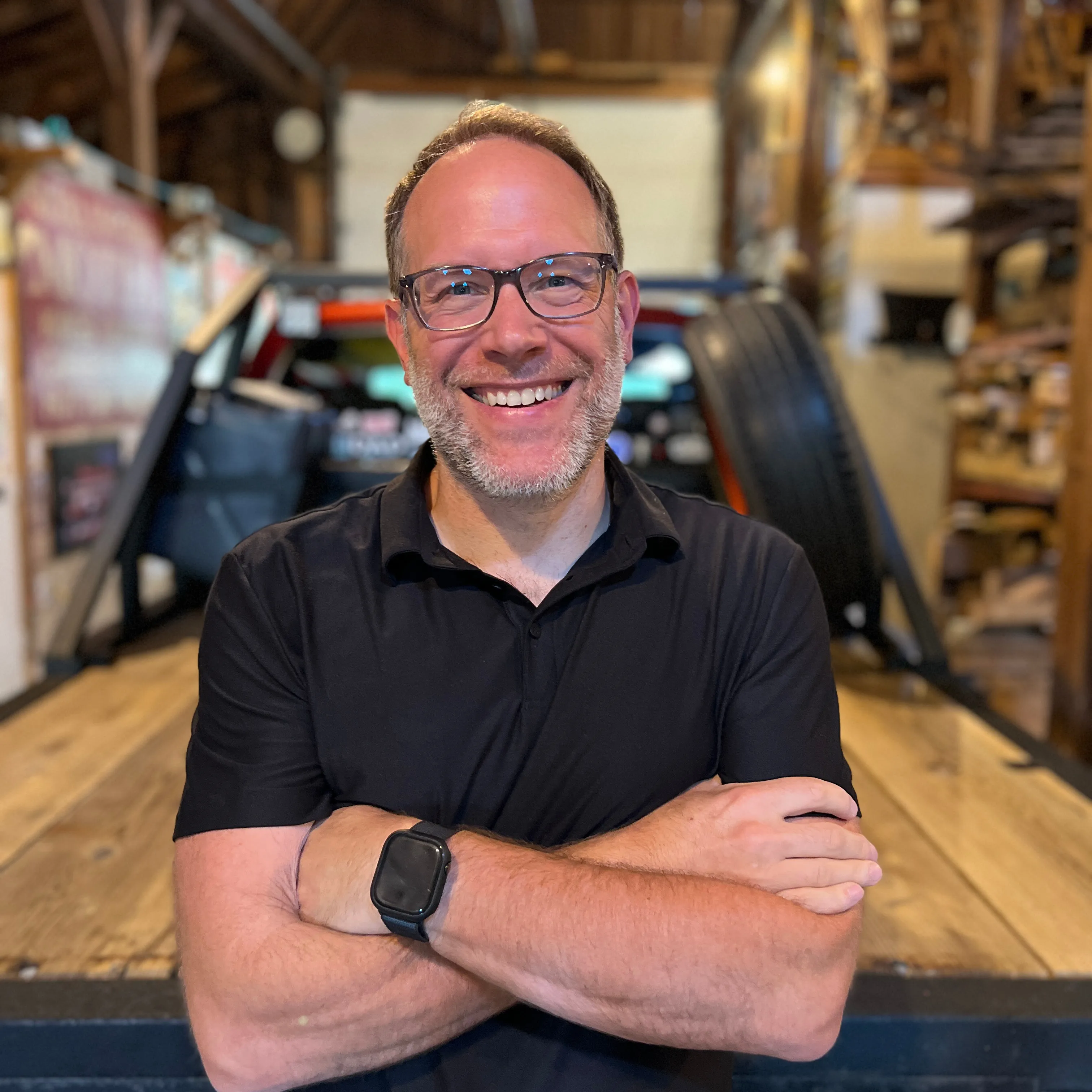 Josh Cowen, Guide MKTG, specializing in Marketing Strategy & Consulting, from Zeeland, Michigan, head-and-shoulders portrait of a smiling man with short brown hair and a close-cropped salt-and-pepper beard wearing glasses and a black short-sleeve collared shirt with arms crossed, standing in front of a blurred rustic workshop interior with a wooden work surface.