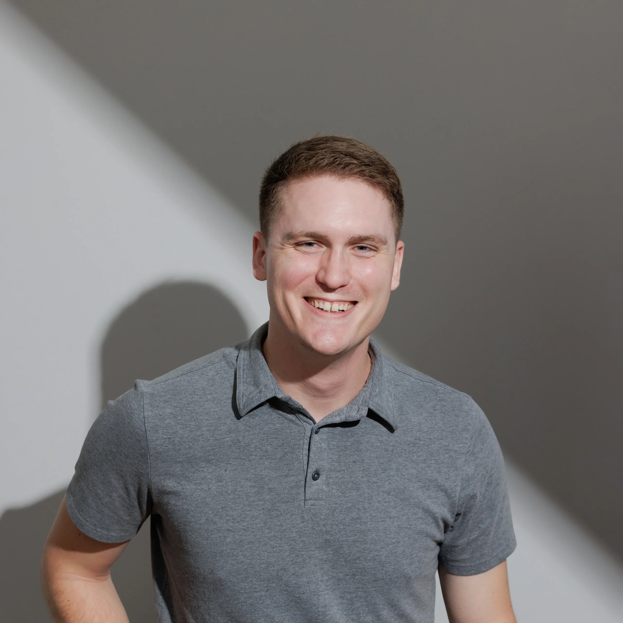 Jordan Ryskamp, Clarity Messaging, specializing in Web Strategy, from Virginia Beach, Virginia, head-and-shoulders portrait of a smiling man with short light brown hair and a light complexion, wearing a gray polo shirt, against a neutral gray wall with a diagonal band of light and soft shadow.
