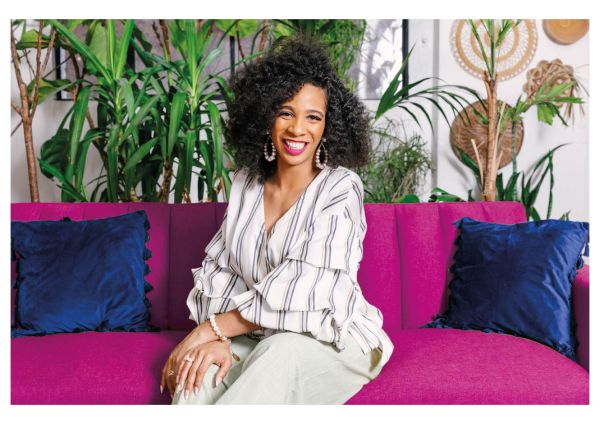 Aleya Harris, Aleya Harris & Spark the Stage, specializing in Public Speaking, from Burbank, California, is shown in a head-and-shoulders seated portrait as a Black woman with voluminous dark curly hair, wearing a white blouse with navy stripes and light trousers, pearl hoop earrings and bracelets, smiling on a magenta sofa with navy pillows against a backdrop of green potted plants.