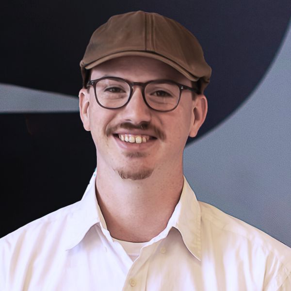 Jon Logan, Greenlit Growth Strategies, specializing in Strategic Business Growth Advisor, from Columbia, South Carolina, head-and-shoulders portrait of a smiling man with short light brown hair, a trimmed mustache and goatee, wearing round eyeglasses, a brown flat cap and a white collared shirt, against a dark blue abstract background.