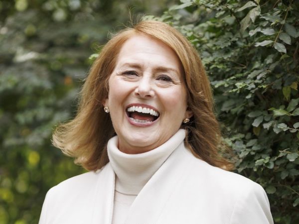 Lisa McGuire, Business Growth Advisor, specializing in Branding & Design, from Alpharetta , Georgia, head-and-shoulders portrait of Lisa McGuire, a woman with shoulder-length light auburn hair and a light complexion, wearing a cream turtleneck and light coat, smiling broadly with teeth visible and small gold stud earrings, photographed outdoors against a softly blurred green leafy background.