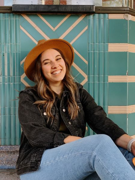 Jill Felty, Felty & Co., specializing in Messaging, from Roseville, California, is shown seated in a three-quarter portrait, smiling with wavy medium-brown shoulder-length hair with lighter ends, wearing a tan wide-brim felt hat, a black denim jacket and light blue jeans, against a turquoise geometric tiled wall.