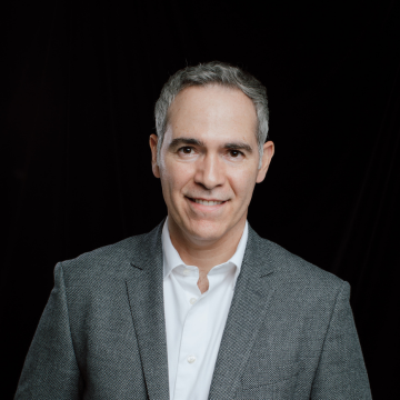Horacio Gutierrez, Capitaltec, specializing in SEO & SEM, from Austin, Texas, head-and-shoulders professional portrait of a clean-shaven man with short gray hair wearing a gray blazer and white button-up shirt, smiling gently and looking at the camera against a plain black background.