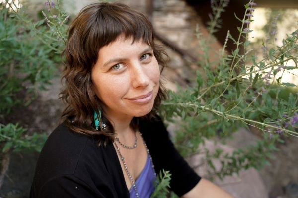 [Paul Zurer], [Magic Words, specializing in Copywriting] from Boulder, Colorado, head-and-shoulders portrait of a woman with shoulder-length wavy brown hair and short bangs, light-medium skin, smiling gently, wearing turquoise drop earrings, a beaded necklace, a purple top and a black cardigan, seated among green leafy plants with small purple flowers against a blurred stone background.