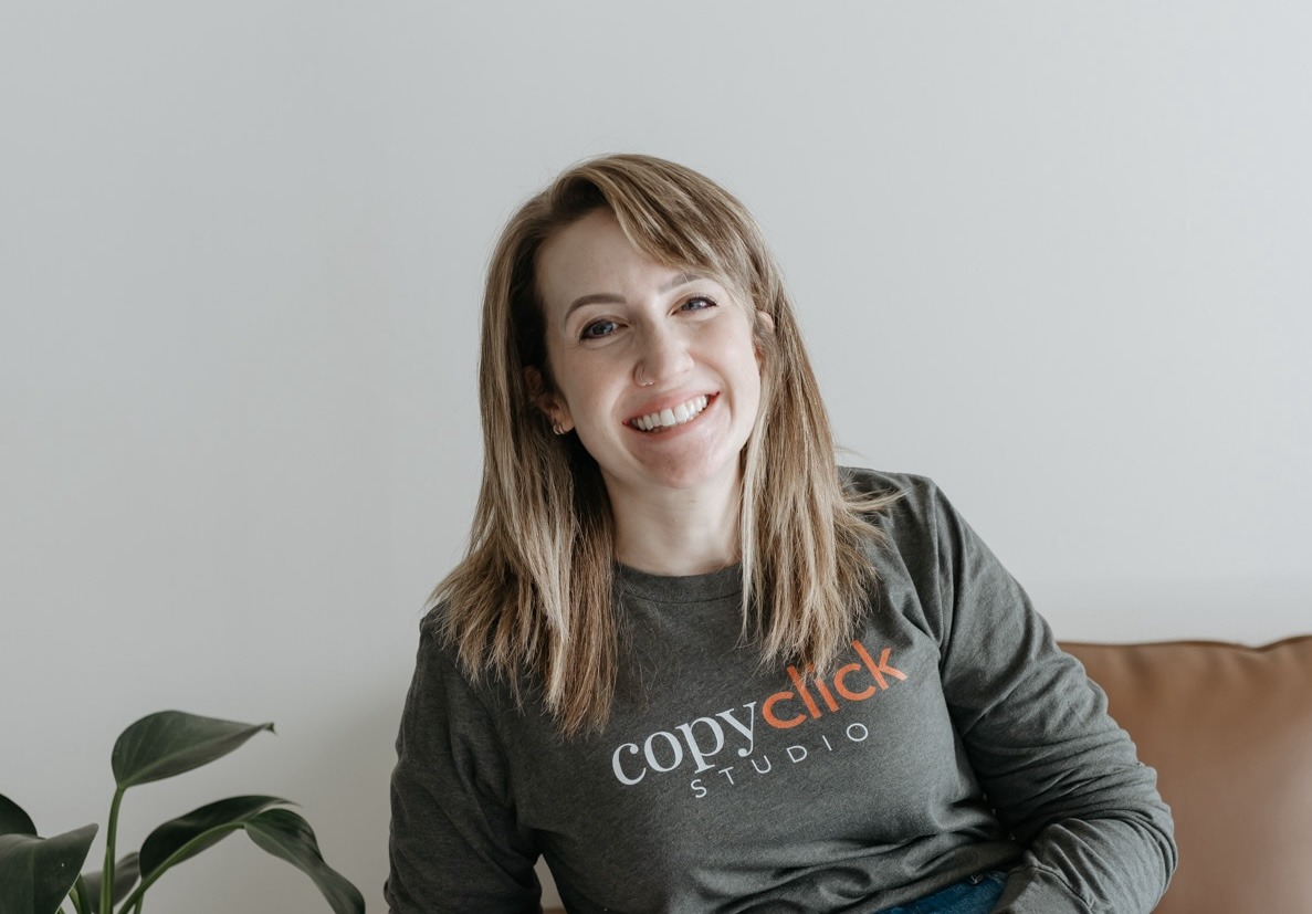Hayley Rissler, CopyClick Studio, specializing in Copywriting, from Crystal Lake, Illinois, head-and-shoulders portrait of a smiling woman with shoulder-length light brown hair and side-swept bangs, wearing a dark gray long-sleeve shirt with an orange-and-white CopyClick Studio logo, seated on a brown leather couch against a light gray wall with a small green houseplant at her left.