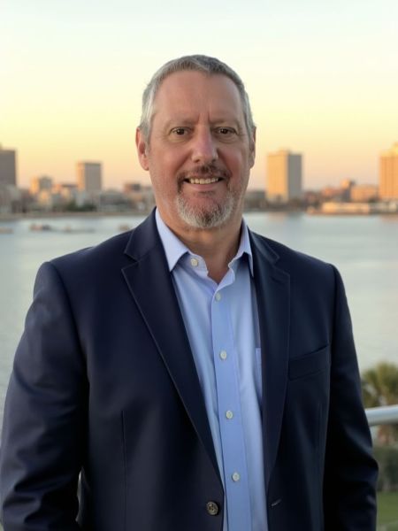 Chuck Boyce, Cibi Creative, specializing in AI Systems Expert, from Mount Pleasant, South Carolina, head-and-shoulders portrait of a middle-aged white man with short salt-and-pepper hair and a trimmed salt-and-pepper beard, wearing a navy blazer and light blue button-down shirt, smiling and standing outdoors on a waterfront with a softly blurred city skyline at sunset.