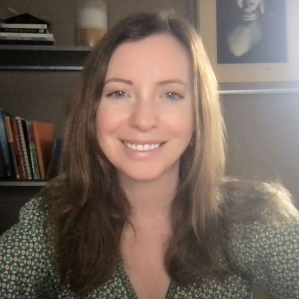 Naomi Catalina, Brand Flow, specializing in Branding & Design, from SANTA ROSA, California, shown in a head-and-shoulders portrait as a smiling woman with long light brown hair worn loose, wearing a green patterned blouse, seated in a bright home office with a bookshelf and framed artwork visible behind her.