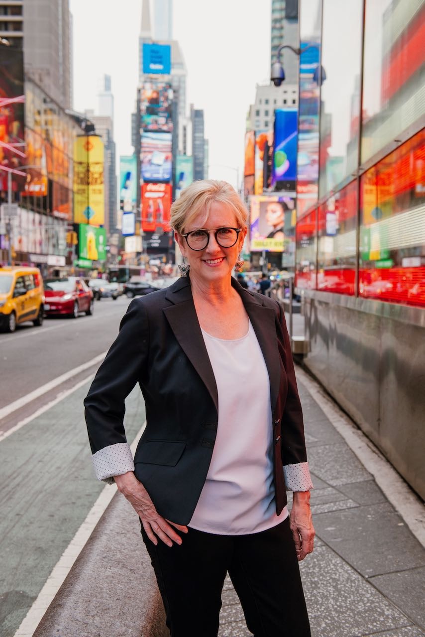 Elaine Acker, Sparx360, specializing in Content Marketing, from Lakeway, Texas, head-and-shoulders portrait of a smiling woman with short blonde hair and black round glasses, wearing a black blazer with rolled polka-dot cuffs over a white blouse, standing with one hand on her hip on a busy city sidewalk with colorful electronic billboards and glass reflections behind her.