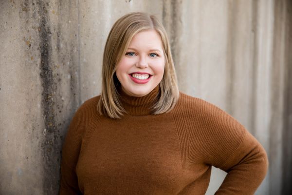 Katelyn Soults, Illuminated, specializing in StoryBrand & HubSpot Agency, from Des Moines, Iowa, head-and-shoulders portrait of a smiling woman with straight shoulder-length blonde hair wearing a brown ribbed turtleneck sweater, leaning slightly against a textured light-gray concrete wall.