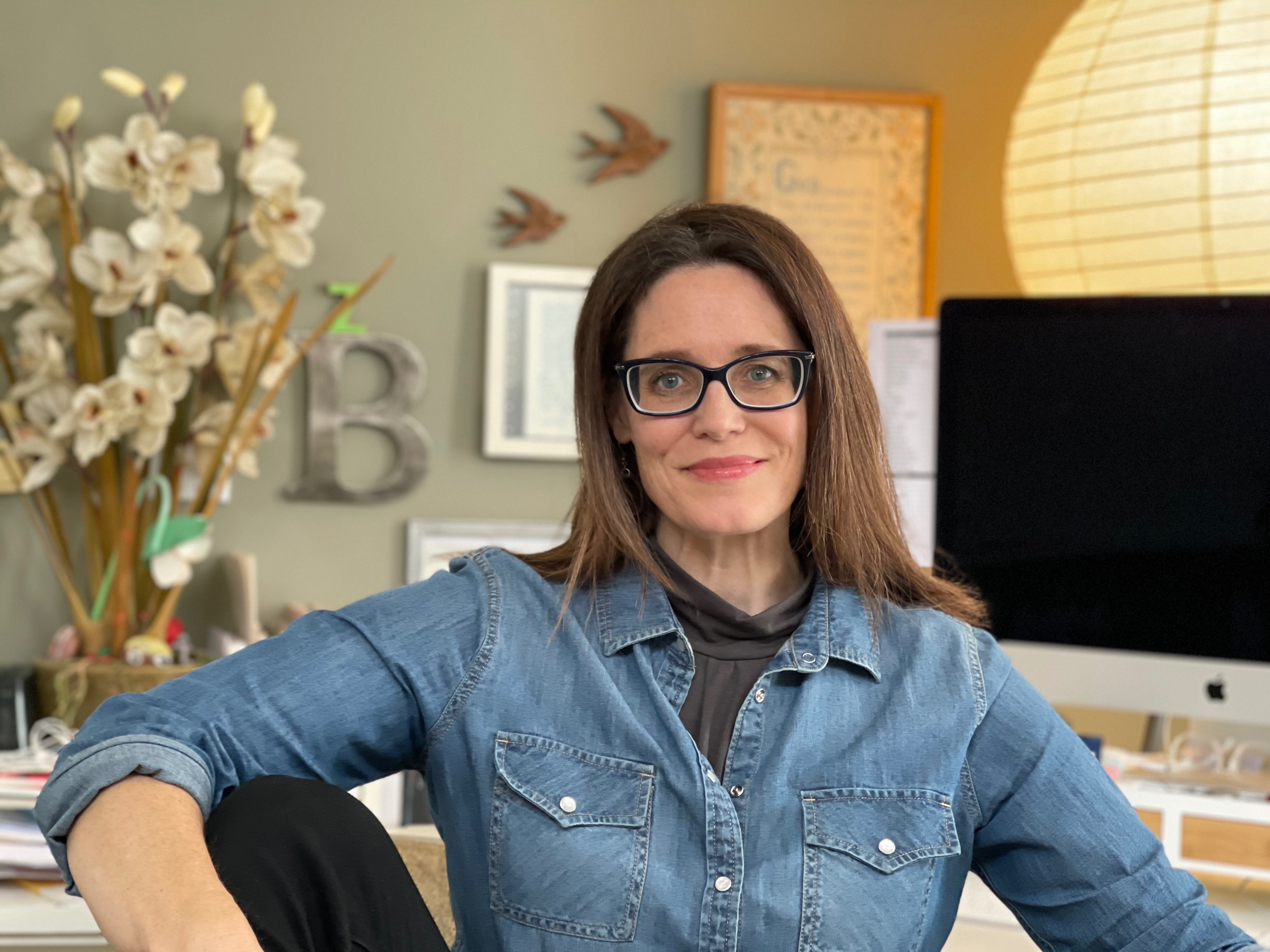 Becky Martin, Guide MKTG, specializing in Marketing Strategy & Consulting, from Zeeland, Michigan, head-and-shoulders portrait of a smiling woman with long straight brown hair wearing black rectangular glasses and a blue denim button-up shirt, seated in a bright home office with a computer monitor and pale floral decor visible behind her.