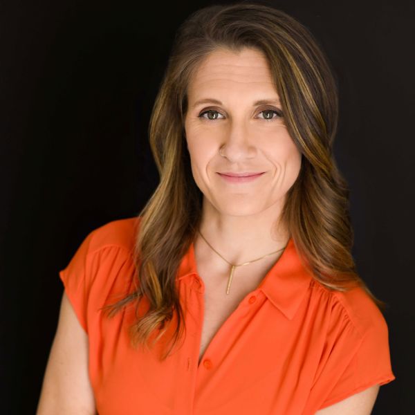 Christine Hehre Tierney, junebird creative, specializing in Marketing Strategy & Consulting, from St. Paul, Minnesota — head-and-shoulders portrait of a fair-skinned woman with shoulder-length brown hair with lighter highlights styled in soft waves, wearing an orange short-sleeve collared blouse, a small nose hoop and a delicate gold vertical bar necklace, smiling gently at the camera against a solid black background.