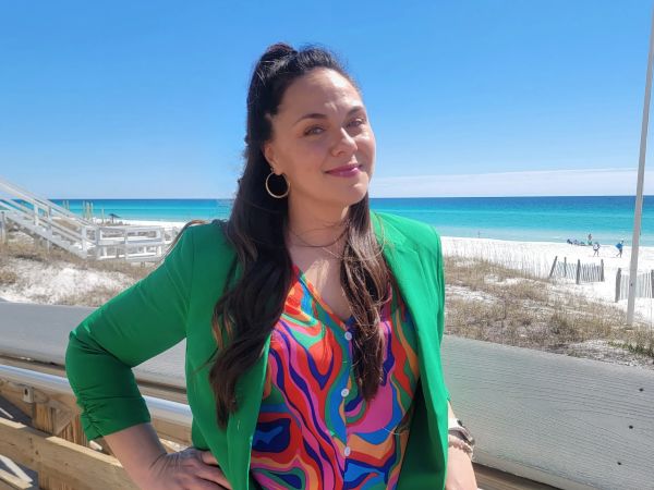 Erica Jones, The Virtual Jones, specializing in Project Management, from Oklahoma City, Oklahoma, head-and-shoulders portrait of a woman with long dark brown hair worn half-up in a ponytail, wearing gold hoop earrings and a bright green blazer over a multicolored patterned blouse, smiling and leaning on a wooden boardwalk railing with white sand and turquoise ocean visible behind her.