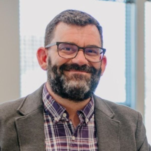 [ Dana Casto ], [ Whispr Strategic Marketing, specializing in Marketing Strategy & Consulting ] from Columbus, Ohio, head-and-shoulders portrait of a smiling man with short dark hair and a salt-and-pepper full beard, wearing dark rectangular glasses, a gray blazer over a purple plaid button-down shirt, photographed indoors in a bright office with a softly lit window background.