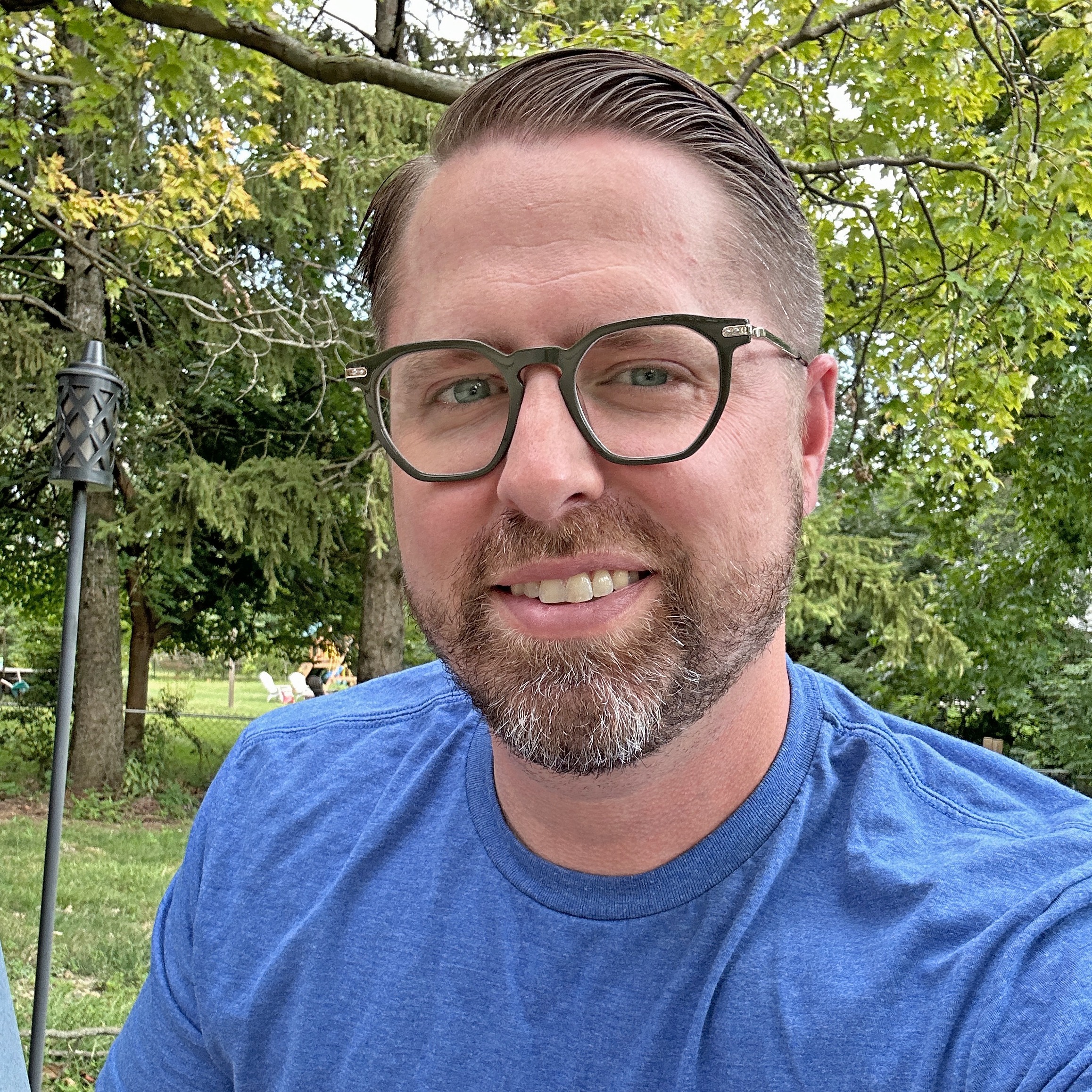 Joe Garrison, Moonflower Marketing, specializing in Marketing Strategy & Consulting, from Indianapolis, Indiana, head-and-shoulders portrait of a man with short light brown hair combed back, a short salt-and-pepper beard, wearing dark olive rectangular glasses and a blue crew-neck T-shirt, smiling while seated outdoors against a backdrop of green trees.