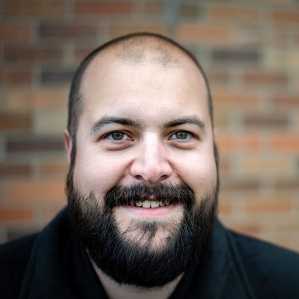 Kellee Carroll, Prairie Giraffe, specializing in SEO & SEM from Gillette, Wyoming, smiling head-and-shoulders portrait of a person with a closely shaved head, full dark beard and mustache, green-hazel eyes, wearing a dark jacket, posed against a softly blurred brick wall.