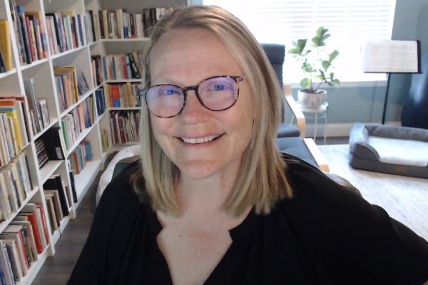 Kelley Hartnett, Tall Tree Collective, specializing in Copywriting, from St. Louis-ish, Missouri — head-and-shoulders portrait of a smiling woman with straight shoulder-length blonde hair wearing tortoiseshell glasses and a black top, seated in a bright home office with a wall of bookshelves, a potted plant by a window, and a dog bed visible.