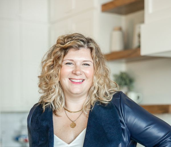 Lauren Cockerell, Kwedar & Co., specializing in Public Relations & Communications, from Fort Worth, Texas, head-and-shoulders portrait of a smiling woman with shoulder-length blonde curly hair wearing a navy jacket over a white top and layered gold necklaces, posed in a bright kitchen with white cabinets and wooden shelves.