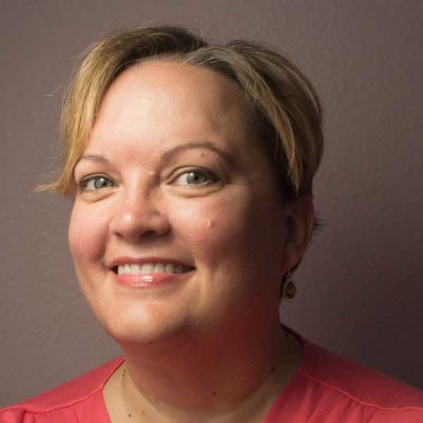 Elizabeth Hahn, Cow and Rooster, specializing in Web Development, from Soulsbyville, California, head-and-shoulders portrait of a smiling woman with short blonde hair swept to the side, wearing a coral blouse and teardrop earrings, photographed against a muted mauve wall.