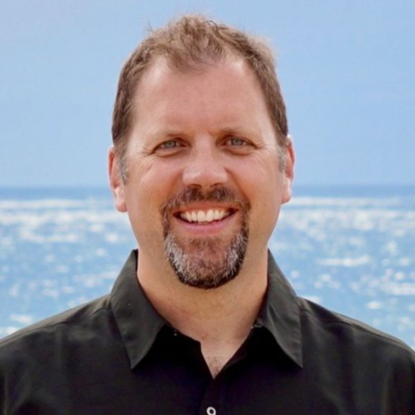 Mike Ruman, Local Growth, specializing in Local & Community Marketing from Colorado Springs, Colorado, is depicted in a head-and-shoulders portrait as a smiling middle-aged man with short brown hair and a trimmed salt-and-pepper goatee, wearing a dark collared shirt against a sunlit ocean background.