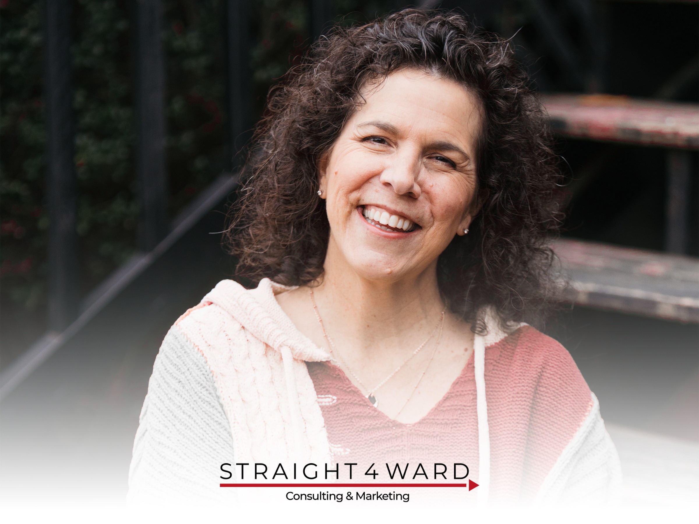 Cassandra Egli, Straight4Ward Consulting & Marketing, specializing in E-commerce Development, from Perrysburg, Ohio, head-and-shoulders portrait of a smiling woman with shoulder-length curly dark brown hair with touches of gray, light-to-medium skin, wearing small stud earrings, layered necklaces, a rust-red knit top and cream cable-knit hoodie, seated on outdoor metal steps with a blurred dark railing and foliage background.