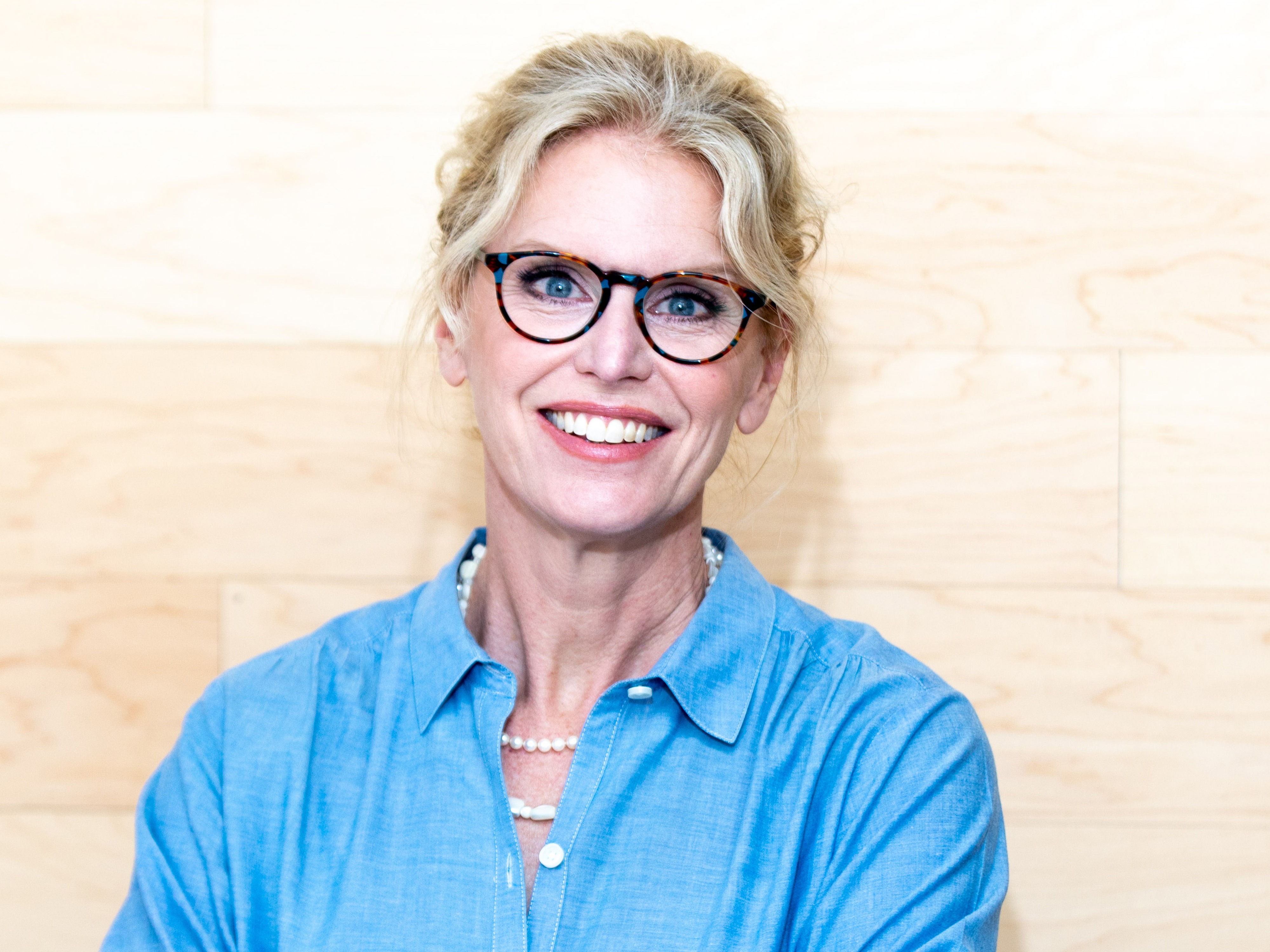 Chris Shull, Heyday Web Media, specializing in Marketing Strategy & Consulting, from Marietta, Georgia, head-and-shoulders portrait of a smiling woman with blonde hair in a loose updo with face-framing strands, wearing tortoiseshell glasses, a light blue button-up shirt and a pearl necklace, posed against a light wood-paneled background.