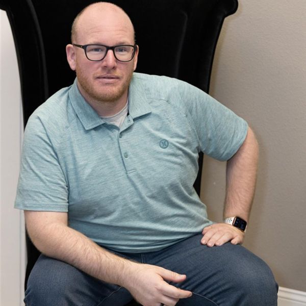 Michael Brant, MRB Media, specializing in AI & Automation, from San Diego, California, head-and-shoulders-to-waist professional portrait of a fair-skinned, bald man with a closely trimmed red beard and black rectangular glasses, wearing a light teal polo over a light undershirt and dark jeans, smiling slightly while seated in a high-back black chair against a neutral beige wall.